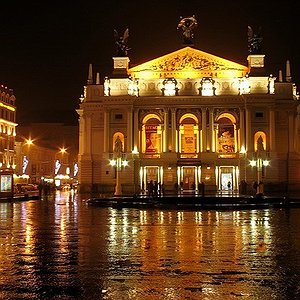 Яскраво освітлений фасад Оперного театру, золоті вогні якого відбиваються на мокрій бруківці